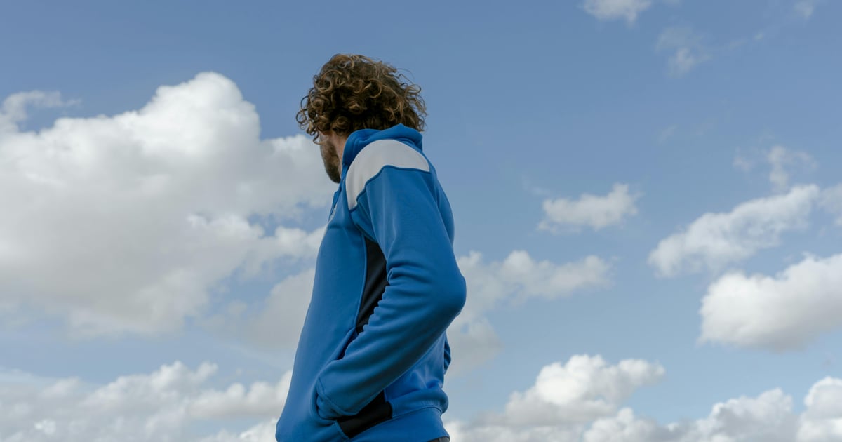 A man with curly hair stands outdoors in a blue jacket, against a cloudy sky.