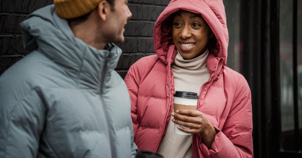 Cheerful multiracial couple in warm clothes with tasty takeaway hot coffee looking at each other while standing near building on street